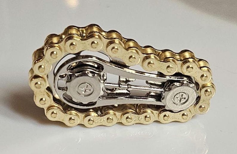 Fidget: A gold-colored bicycle chain link design with silver-colored mechanical components. The chain is arranged in a loop around the silver-colored parts. The object is on a white surface.