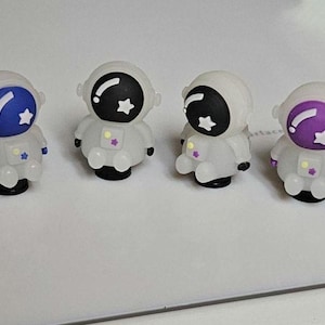 May include: Six small astronaut figurines in a row. Each astronaut is white with a colored visor and star details. The visors are blue, black, and purple. The figurines have black bases.