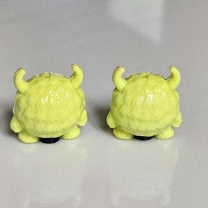以下が含まれることがあります： 小さな黄色のモンスター型のイヤリングのペア。各イヤリングは丸みを帯びたボディ、2つの湾曲した角、黒いベースを備えています。イヤリングは質感のある毛皮のような外観で、白い背景に配置されています。