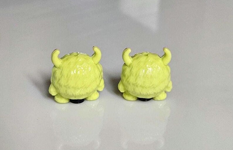 以下が含まれることがあります： 小さな黄色のモンスター型のイヤリングのペア。各イヤリングは丸みを帯びたボディ、2つの湾曲した角、黒いベースを備えています。イヤリングは質感のある毛皮のような外観で、白い背景に配置されています。