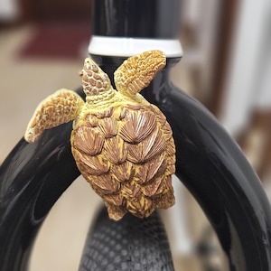 May include: A detailed, bronze-colored turtle figurine attached to a black bicycle fork. The turtle has textured details on its shell and flippers. The bicycle tire is black with a textured pattern. The text "SE OTW OFF THE WALL" is visible on the bicycle.