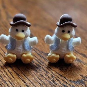 May include: Two small, white penguin figurines with yellow feet and beaks, wearing blue vests and brown bowler hats. The penguins are standing on a wooden surface.