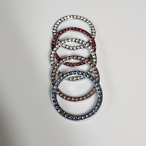 May include: A vertical stack of five circular metal rings, each featuring a single row of embedded crystals. The rings display alternating colors including clear, deep red, light pink, and blue, all set within a polished silver-tone metal frame.