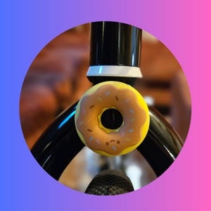 May include: A yellow donut-shaped bicycle bell with brown frosting and sprinkles. The bell is mounted on a black bicycle frame, set against a blurred background. The bell has a small, smiling face.