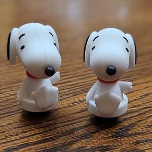May include: Two small white Snoopy figurines with black ears and spots, wearing red collars. The figures are sitting on a wooden surface. The figurines are facing the viewer.