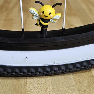 May include: A yellow and black bee-shaped tire valve cap is attached to a black bicycle tire. The bee has white and yellow wings and black antennae. The tire has a white sidewall and a black tread.