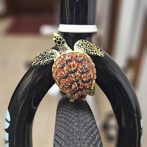 May include: Close-up of a black bicycle fork with a textured tire. A decorative turtle figurine with brown and yellow details is attached to the fork. The fork has a sticker that says "SE OTW OFF THE WALL".