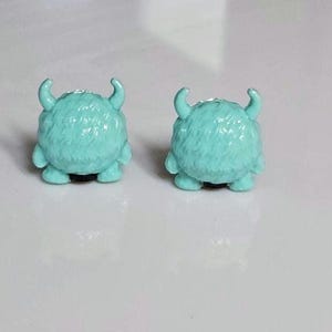 以下が含まれることがあります： ターコイズ色のモンスター型スタッドピアス。各ピアスは、丸く毛むくじゃらの体、2つの湾曲した角、小さな足が特徴です。ピアスは無地の白い背景に配置されています。