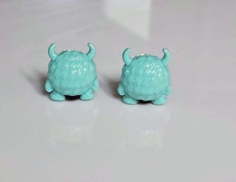 以下が含まれることがあります： ターコイズ色のモンスター型スタッドピアス。各ピアスは、丸く毛むくじゃらの体、2つの湾曲した角、小さな足が特徴です。ピアスは無地の白い背景に配置されています。