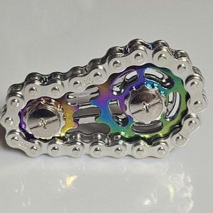 A silver-colored fidget spinner in the shape of a bicycle chain. The center features a rainbow-colored design with two silver screws. This unique spinner is designed to relieve stress and improve focus.