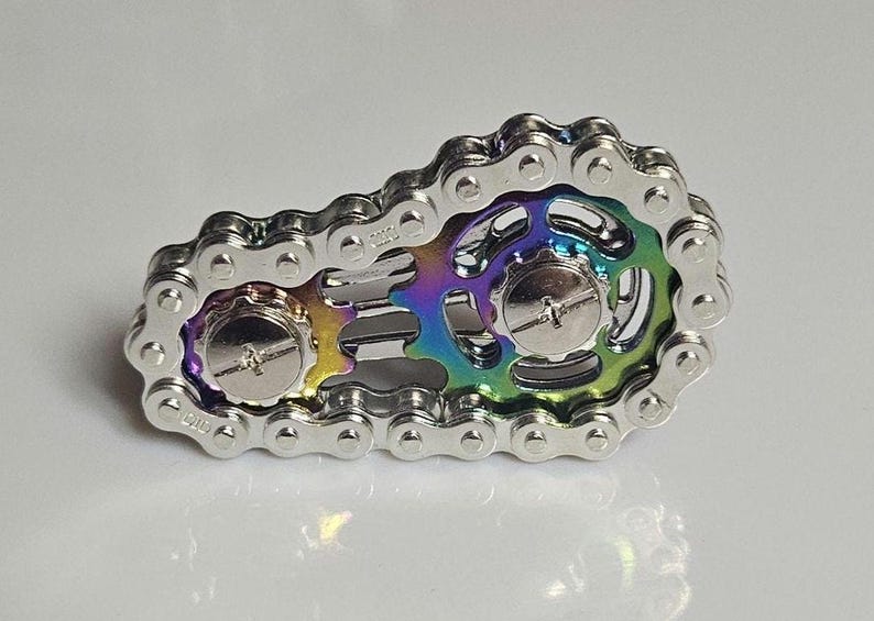 A silver-colored fidget spinner in the shape of a bicycle chain. The center features a rainbow-colored design with two silver screws. This unique spinner is designed to relieve stress and improve focus.