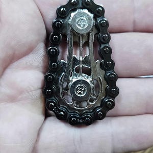A silver and black metal fidget featuring a bicycle chain design. The pendant has a central gear and two smaller gears, all connected by a black chain. The design is intricate and detailed, with a focus on mechanical elements.