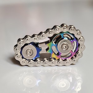 Może przedstawiać: Srebrny fidget spinner w kształcie łańcucha rowerowego. Centralne koła zębate mają tęczowe wykończenie z niebieskimi i zielonymi odcieniami. Spinner jest przeznaczony do trzymania i obracania palcami.