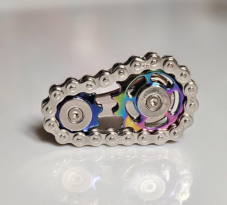 Może przedstawiać: Srebrny fidget spinner w kształcie łańcucha rowerowego. Centralne koła zębate mają tęczowe wykończenie z niebieskimi i zielonymi odcieniami. Spinner jest przeznaczony do trzymania i obracania palcami.