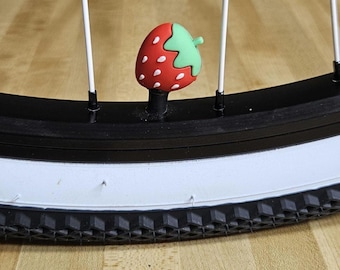 May include: A red and white strawberry-shaped valve cap on a bicycle tire. The tire is black with a white sidewall and a black tread. The strawberry cap has a green stem and white seeds.