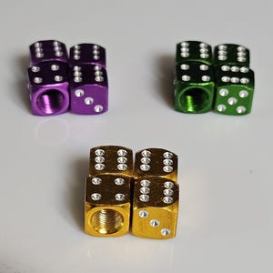 May include: Three sets of dice-shaped tire valve stem caps. One set is purple, one is green, and the other is gold. Each cap has white dots representing the numbers on a die. The caps are made of metal.