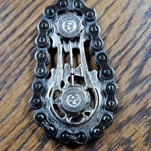 Może przedstawiać: Srebrzysta metalowa zabawka typu fidget, w kształcie łańcucha i k&oacute;ł zębatych roweru. Zabawka ma szczeg&oacute;łowy design z zazębiającymi się ogniwami łańcucha i okrągłymi kołami zębatymi, tworząc mechaniczną estetykę. Tło to drewniana powierzchnia.