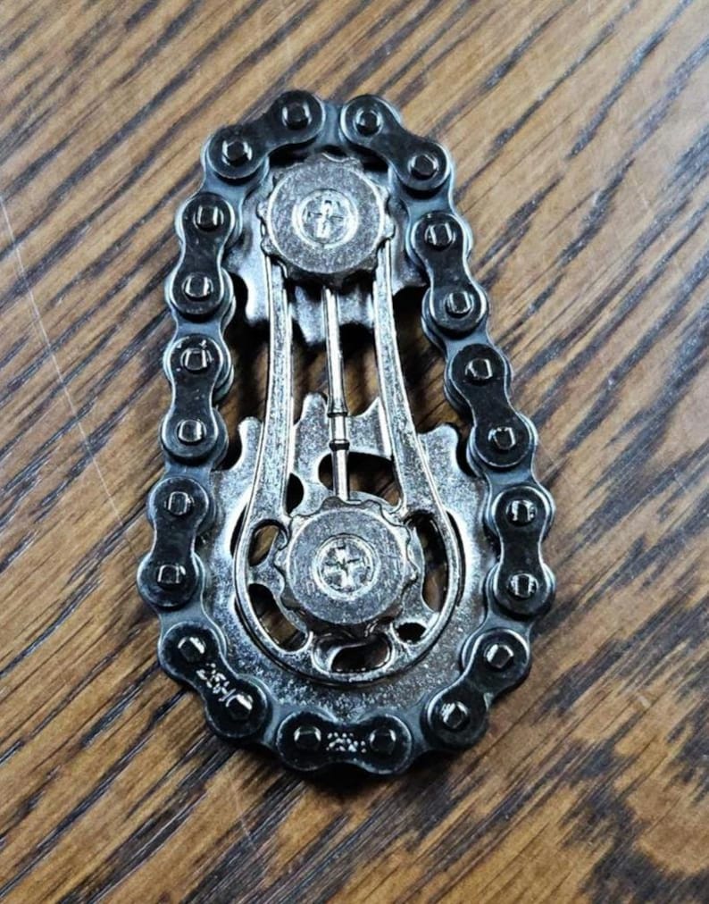 Może przedstawiać: Srebrzysta metalowa zabawka typu fidget, w kształcie łańcucha i k&oacute;ł zębatych roweru. Zabawka ma szczeg&oacute;łowy design z zazębiającymi się ogniwami łańcucha i okrągłymi kołami zębatymi, tworząc mechaniczną estetykę. Tło to drewniana powierzchnia.