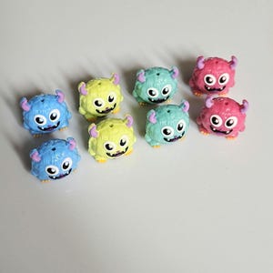 以下が含まれることがあります： 8体の小さなカラフルなモンスターのフィギュア。 モンスターは青、黄、緑、ピンクの色で、それぞれ2本の角、大きな目、笑顔の口があります。 明るい色の表面に配置されています。 楽しくてカラフルなコレクションです。