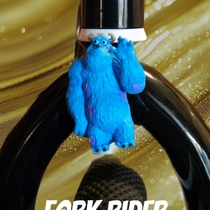 May include: A blue monster figurine with purple accents, positioned on a black object. The text "FORK RIDER" is displayed in white, with the "Bazaart" logo in the bottom right corner. The background is a blurred golden color.
