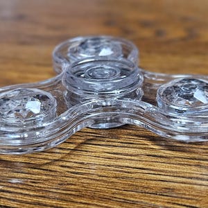 May include: Clear, transparent fidget spinner with a three-lobed design. Each lobe features a faceted, gem-like embellishment. The spinner is made of a clear material and rests on a wooden surface.