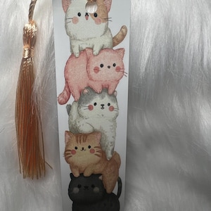 May include: A rectangular bookmark featuring a stack of six cartoon cats in various colors: white, pink, gray, orange, and black. The bookmark has a gold tassel attached to the top.