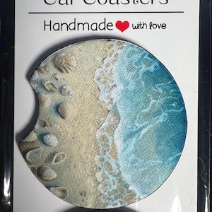 May include: A round, beige and blue car coaster with a beach scene. The coaster features a sandy beach with seashells and a blue ocean wave crashing on the shore. The text "Handmade with love" is printed on the coaster.