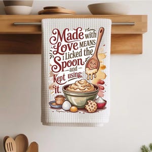 May include: White waffle-weave kitchen towel with a colorful illustration and text. The illustration features a wooden spoon with a smiling face, a bowl of cream, and the text "Made with Love Means I Licked the Spoon and Kept using it."