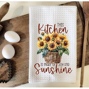 Puede incluir: Toalla de cocina blanca con una cesta marrón de girasoles y el texto "This Kitchen is made of Joy and Sunshine".
