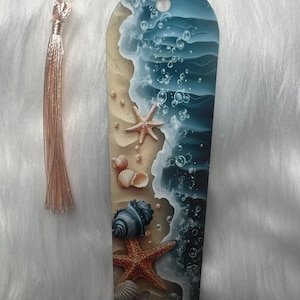 Puede incluir: Marcapáginas decorativo con diseño de playa. Representa una playa arenosa, olas del océano, estrellas de mar, conchas y burbujas. El marcapáginas tiene una borla. La paleta de colores incluye tonos azules, beige y naranja.