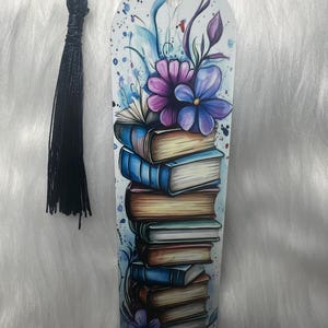 May include: A decorative bookmark featuring a stack of books with blue and gold covers, adorned with purple and blue flowers and watercolor accents. A black tassel hangs from the top. The bookmark is a unique accessory for book lovers.
