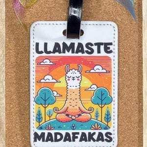 Op de afbeelding: Wit leren bagagelabel met een kleurrijke illustratie van een mediterende lama met de tekst "LLAMASTE MADAFAKAS".