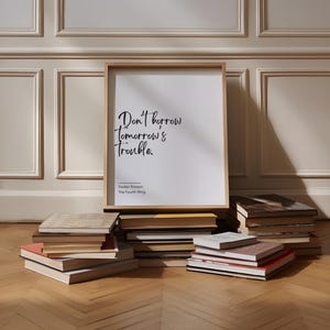 May include: A framed print with a white background and black text that reads "Don't borrow tomorrow's trouble." The text is in a handwritten style. Below the text is the name "Kaden Ripson" and "The Fourth Wing". The print is leaning against a stack of books on a wooden floor.