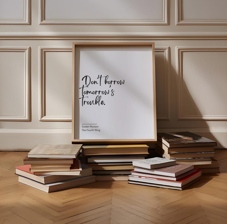 May include: A framed print with a white background and black text that reads "Don't borrow tomorrow's trouble." The text is in a handwritten style. Below the text is the name "Kaden Ripson" and "The Fourth Wing". The print is leaning against a stack of books on a wooden floor.