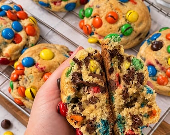 Puede incluir: Una galleta grande y gruesa partida por la mitad que revela un interior suave y pastoso relleno de chispas de chocolate negro y dulces de chocolate de colores. Varias galletas enteras con coberturas similares descansan en una rejilla.