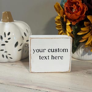 May include: A rectangular wooden block sign with a distressed white finish and dark brown edges. The front face features the black printed text "your custom text here" centered in a simple serif font, showcasing a customizable decorative piece.