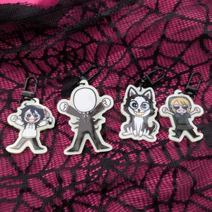 Puede incluir: Cuatro llaveros de personajes de dibujos animados con clips negros. Los personajes incluyen una figura con la cara en blanco, un perro sonriente, una figura con un cuchillo y una figura con ropa verde. Los llaveros están sobre un fondo rosa y negro con telarañas.