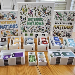 May include: Three white board game boxes titled Notebook Nations with colorful character illustrations, accompanied by several white plastic organizers containing stacks of playing cards, small circular tokens, and game pieces.