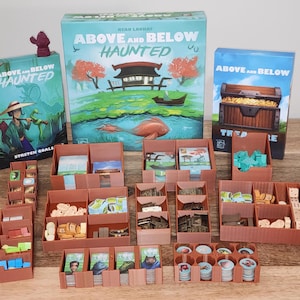 May include: A board game titled "Above and Below Haunted" with various game components. The game boxes feature illustrations of a Japanese-style scene with a boat and a treasure chest. The game components are organized in brown trays.