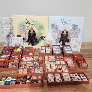 May include: A board game titled "Darwin's Journey" with various game components, including game boxes, cards, and tokens, arranged in orange trays. The game boxes feature illustrations of Charles Darwin.