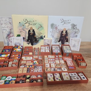 Puede incluir: Un juego de mesa llamado "Darwin's Journey" con varios componentes. El juego incluye cajas, cartas y fichas, dispuestas en bandejas naranjas. Las cajas del juego presentan ilustraciones de Charles Darwin.