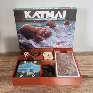 May include: A board game box titled "Katmai: The Bears of Brooks River" featuring a brown bear catching a fish in a river. The game box sits above an orange organizer with game pieces and cards.