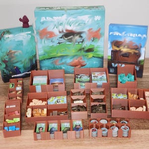 May include: A board game with three boxes and several brown storage containers filled with game pieces. The boxes have colorful abstract art. The storage containers hold cards, tokens, and other game components.