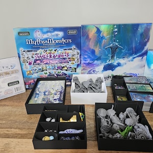 May include: A board game collection featuring the Avalon Myths & Monsters expansion box, the Iron Forged upgrade pack, and a large game box with a sword illustration. Includes black and white plastic organizers filled with grey miniature figures.