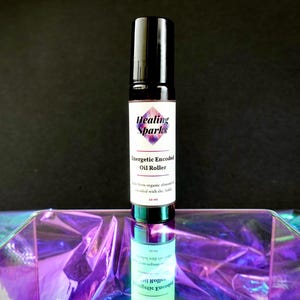 Könnte beinhalten: Eine Roll-on-Flasche mit schwarzem Deckel und weißem Etikett mit der Aufschrift "Healing Sparks" und "Energetic Encoded Oil Roller". Die Flasche ist 15 ml groß und steht auf einer reflektierenden, irisierenden Oberfläche.