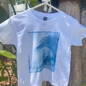 Peut inclure: T-shirt blanc avec une image bleue et blanche d'un surfeur chevauchant une vague.