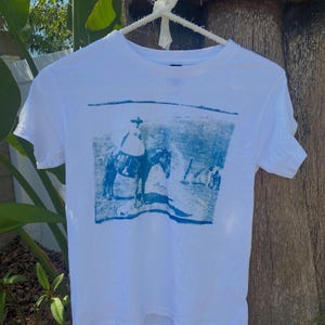 May include: White t-shirt with a blue image of a person riding a horse. The image is a vintage style print.