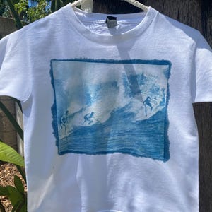 May include: White t-shirt with a blue image of surfers riding waves. The image is framed with a white border.