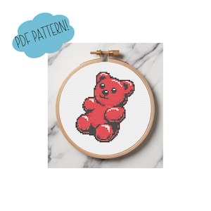 May include: A cross-stitch design of a red gummy bear in a wooden embroidery hoop. The text "PDF PATTERN!" is in a blue speech bubble in the upper left corner. The background is a white marble surface.