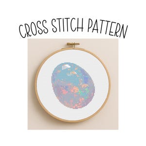 May include: A cross stitch pattern featuring a colorful, oval-shaped design resembling an opal gemstone. The design is set within a wooden embroidery hoop. The words "CROSS STITCH PATTERN" are arched above the design.
